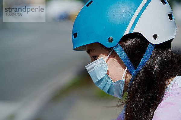 Mädchen in Maske und Helm fahren Kick-Scooter auf der Straße in der Stadt Straße