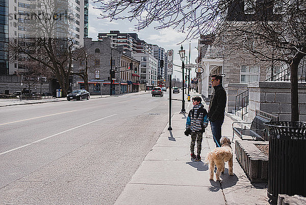 Vater und Sohn mit Hund stehen auf dem Bürgersteig einer ruhigen Stadtstraße.