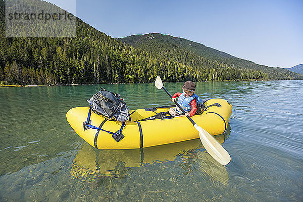 Seitenansicht eines Jungen  der mit einem gelben aufblasbaren Kajak auf einem ruhigen See paddelt.