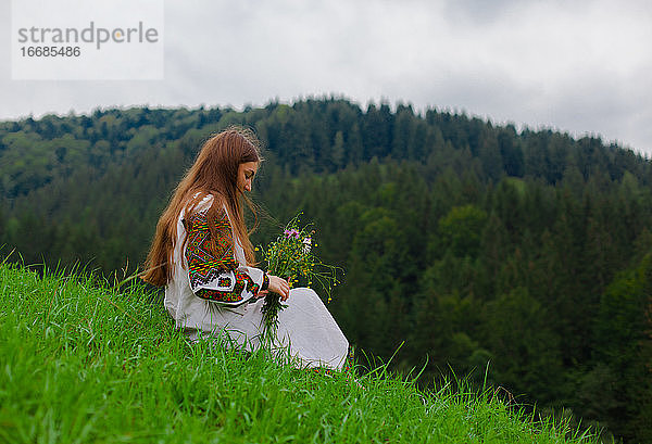 Mädchen in Stickerei mit einem Strauß Wildblumen sitzt auf grünem Gras in den karpatischen Bergen