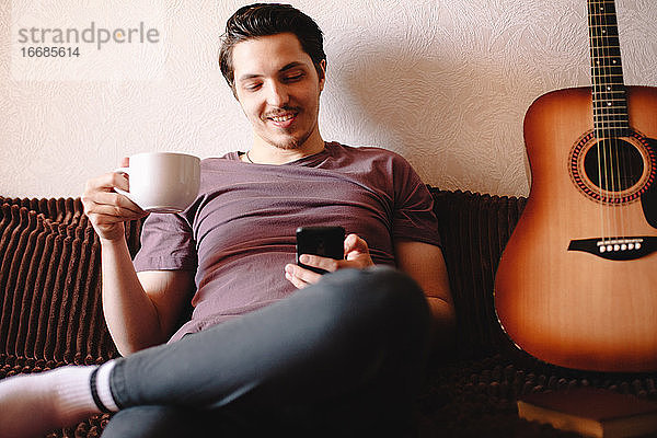 Glücklicher junger Mann  der sein Smartphone benutzt  während er zu Hause Tee trinkt