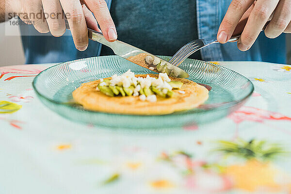 Ein Teller mit Pfannkuchen mit Käse und Avocado steht auf dem Tisch. Ein anonymer Mann isst mit einem bunten Tischtuch