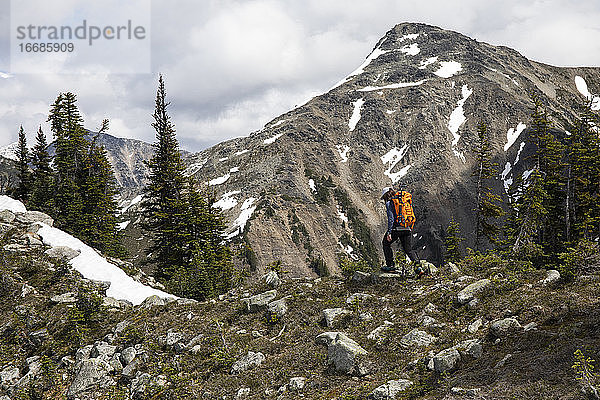 Seitenansicht eines anonymen Reisenden mit Rucksack  der auf einem steinigen  mit Schnee und Wald bedeckten Berghang in British Columbia in Kanada wandert