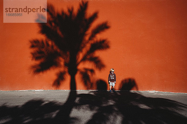 Junge steht vor einer roten Wand mit dem Schatten einer Palme