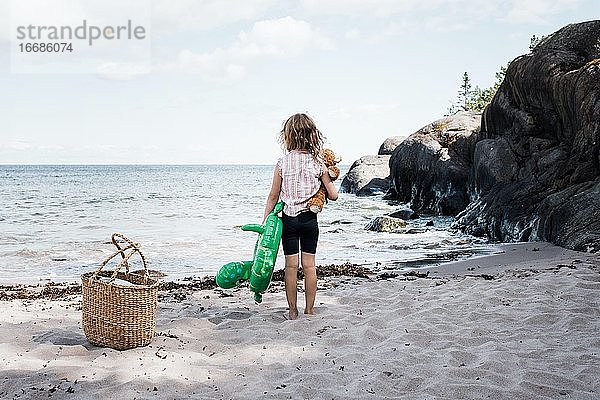 Mädchen stand am Strand mit ihrem Bären und dem Schlauchboot am Meer