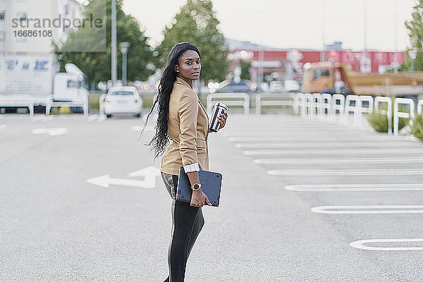 Porträt einer schwarzen Frau in einem braunen Anzug  die einen Laptop und einen Behälter mit Kaffee in der Hand hält und die Straße entlanggeht