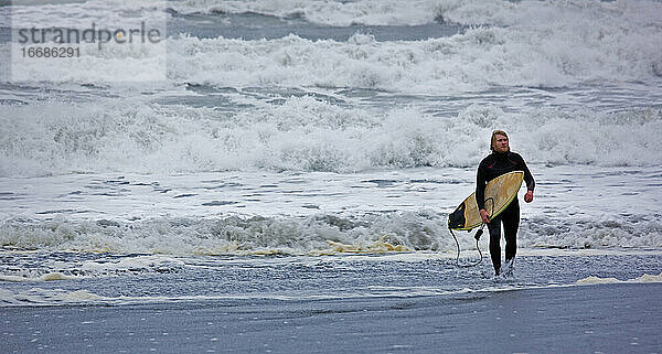 junger Mann geht mit Surfbrett aus dem Wasser in Island