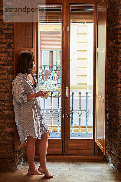 Junge Frau beim Frühstück neben einem Fenster zu Hause.