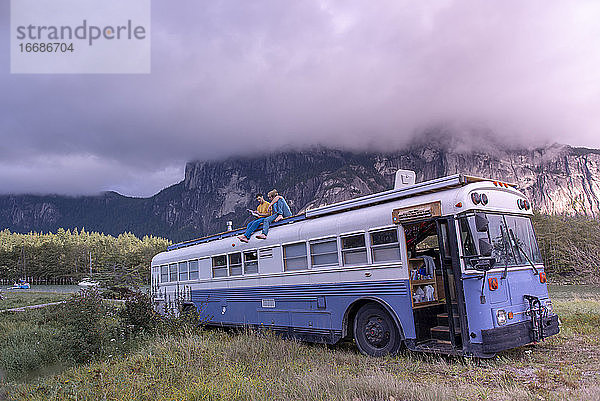 zwei Personen sitzen auf dem Dach eines Schulbusses und lesen ein Buch vor einem Berg