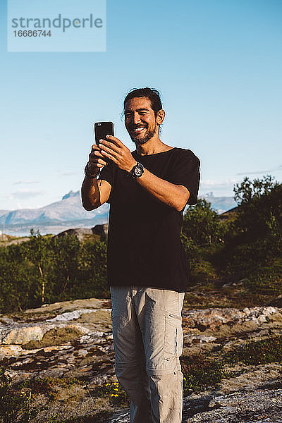 Lächelnder Mann  der ein Foto mit seinem Smartphone auf einem Berggipfel macht