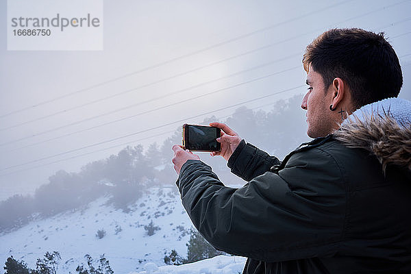 Ein junger lateinamerikanischer Mann macht ein Foto mit seinem Handy  umgeben von Schnee