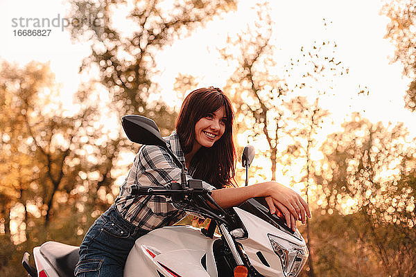 Porträt einer glücklichen jungen Frau  die auf einem Motorrad inmitten von Bäumen sitzt