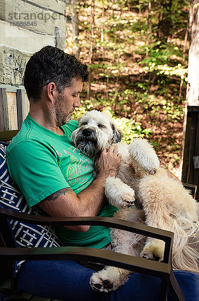 Mann  der einen flauschigen Hund auf seinem Schoß hält  draußen auf einer Terrasse im Wald.