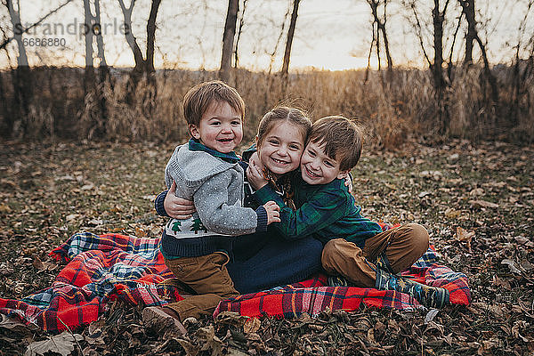 Drei Geschwister sitzen auf einer Decke und umarmen sich und lächeln in die Kamera