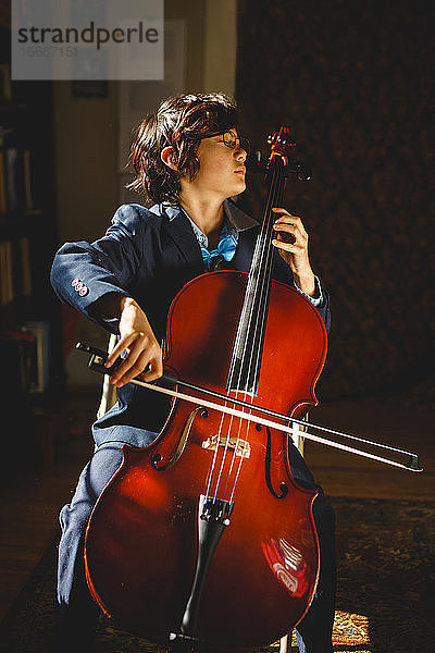 Ein ernster Junge in Anzug und Fliege mit Brille spielt zu Hause Cello