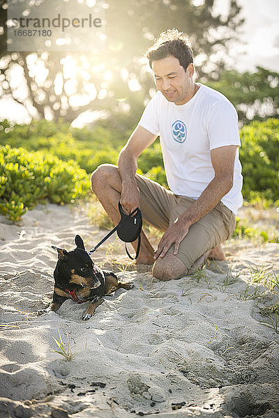 Junger fitter Mann hockt am Strand mit seinem kleinen schwarzen Hund in Hawaii