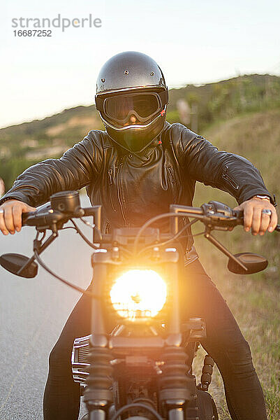 Biker mit Helm sitzt und entspannt auf einem alten Motorrad bei Sonnenuntergang.