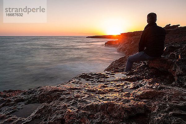 Mann sitzt auf den Felsen am Meer und beobachtet den Sonnenuntergang. Langzeitbelichtung