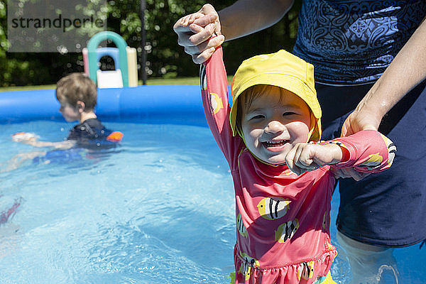 Happy Baby Girl Wearing Yellow Sun Hat Plays in Pool mit Familie