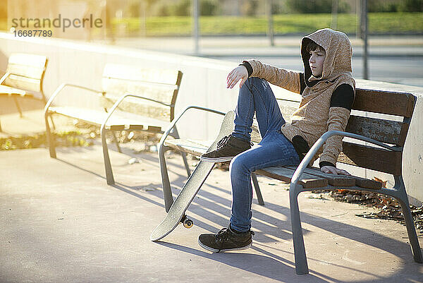 Junger Junge mit Kapuze sitzt auf einer Bank neben seinem Skateboard