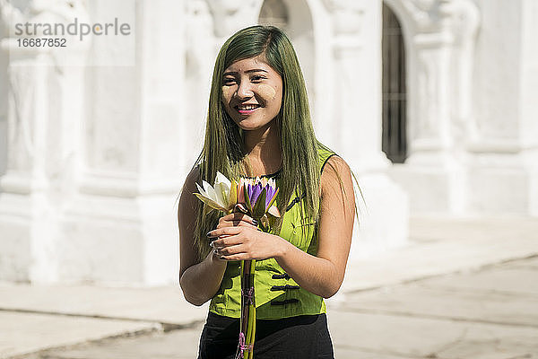 Lächelnde junge Frau in grüner Freizeitkleidung hält Blumen vor der Kuthodaw-Pagode  Mandalay  Myanmar
