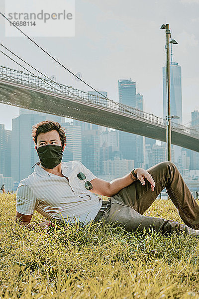 Junger Mann sitzt im Park und trägt eine Gesichtsmaske.