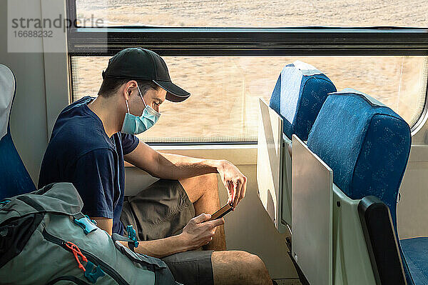 Rucksacktourist mit Gesichtsmaske  der während einer Zugfahrt ein Mobiltelefon benutzt.