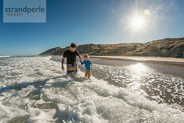 Lächelnder Vater und Kind spielen in den Wellen an einem sonnigen Tag an einem Strand in Neuseeland
