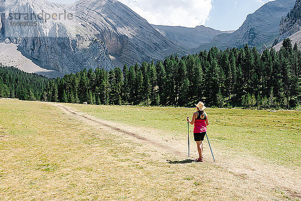 Wanderer Frau trägt Strohhut  Shorts und Rucksack auf dem Weg über eine Ebene zu Fuß mit erstaunlichen Berge im Hintergrund  während genießt die natürliche Umgebung um. Horizontal Foto.