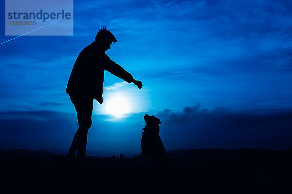 Silhouette eines jungen Mannes  der seinen Hund mit blauem Nachtlicht füttert