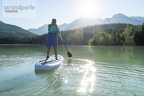 Rückenansicht einer barfuß lebenden Frau in Kleid und Schwimmweste auf einem SUP-Board auf einem ruhigen See an einem sonnigen Sommertag in den Bergen in British Columbia  Kanada