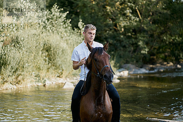 Porträt eines jungen blonden Mannes  der auf einem Pferd über einen Fluss reitet