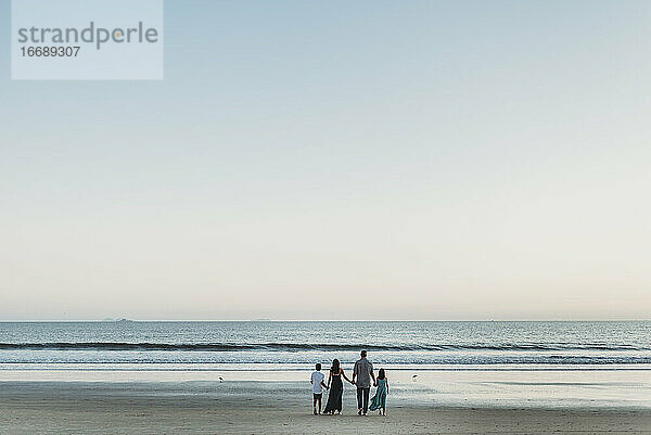Junge Familie steht am Strand und hält sich an den Händen  während sie auf das Meer hinausschaut