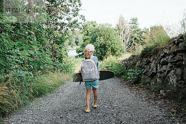 junges Mädchen  das mit einem Skateboard und einem Rucksack einen Feldweg entlang läuft