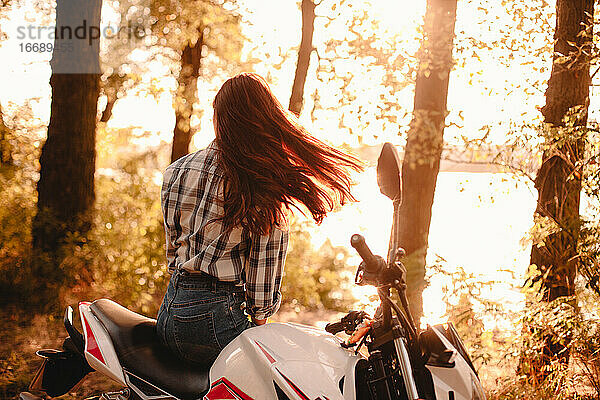 Junge Frau wirft sich entspannt die Haare und sitzt auf einem Motorrad am Fluss