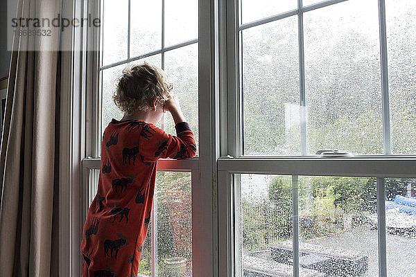 Rear View of Young Boy With Curly Hair Looking Out Rain Covered Window