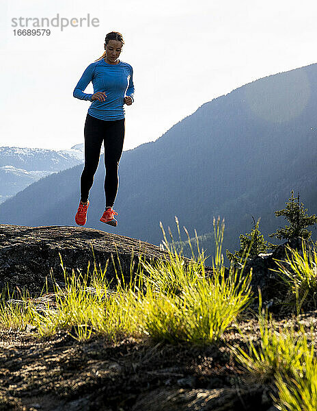 Vorderansicht einer Sportlerin in Sportkleidung  die an einem sonnigen Abend auf dem Lande auf Felsen in der Nähe von Bergen läuft