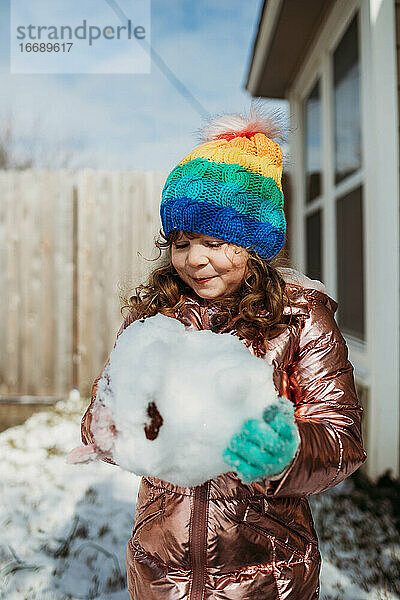 Junges Mädchen mit Regenbogenhut hält riesigen Schneeball im Winter