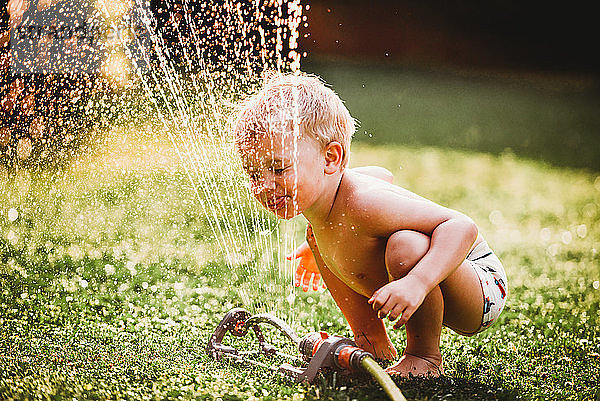 Weißer Junge streckt die Zunge heraus und trinkt Wasser aus dem Rasensprenger im Garten