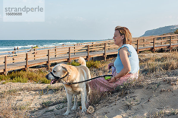 Frau schaut auf das Meer  während sie mit ihrem Hund im Sand sitzt