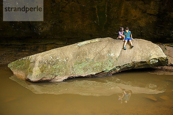 zwei Kinder sitzen auf einem großen Felsbrocken über einem spiegelnden Becken an einer Steinmauer