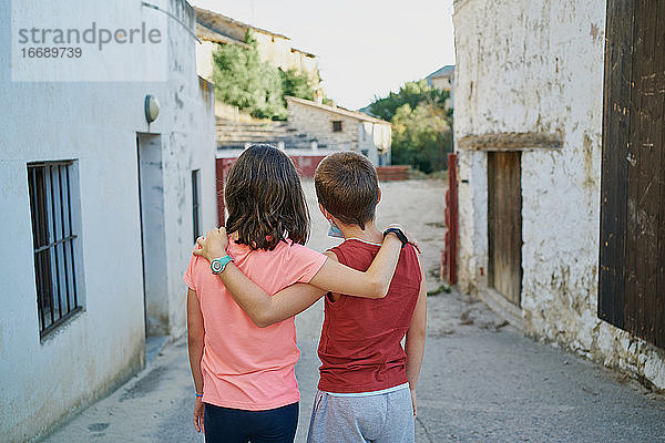 Rückenansicht von Kindern  die in einem spanischen Dorf in Teruel mit einer Stierkampfarena im Hintergrund umarmt die Straße entlanglaufen