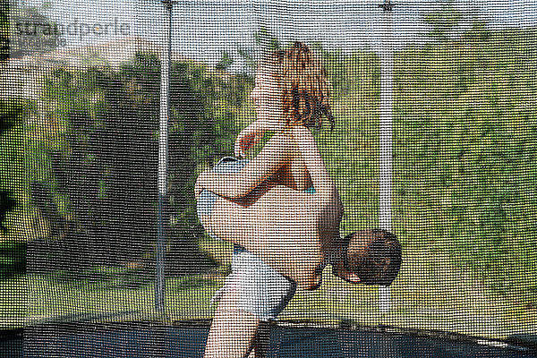Horizontales Foto eines blonden Jungen und eines brünetten Mädchens  die auf einem Trampolin auf einem grünen Spielplatz kämpfen und spielen. Der Teenager und das Kind tragen kurze Jeans und sie sehen glücklich aus  lächelnd