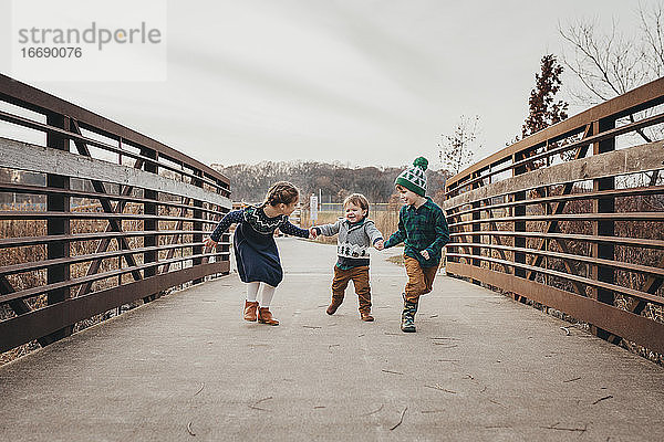 Drei lachende Geschwister  die sich an den Händen halten und auf einer Brücke laufen