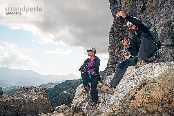 Konzept: Abenteuer. Mann und Frau klettern mit Helm und Klettergurt. Sie ruhen sich auf einem Felsen sitzend aus. Sie unterhalten sich und gestikulieren  während sie auf den Horizont schauen. Via ferrata in den Bergen.