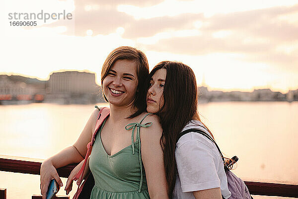 Zwei glückliche Freundinnen stehen auf einer Brücke gegen den Fluss bei Sonnenuntergang
