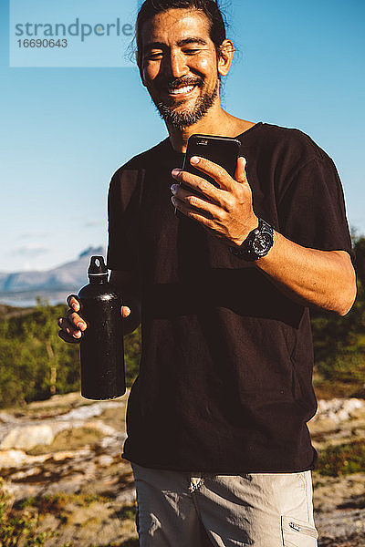 Mann lächelnd hält sein Telefon und eine Wasserflasche in einem sonnigen Tag