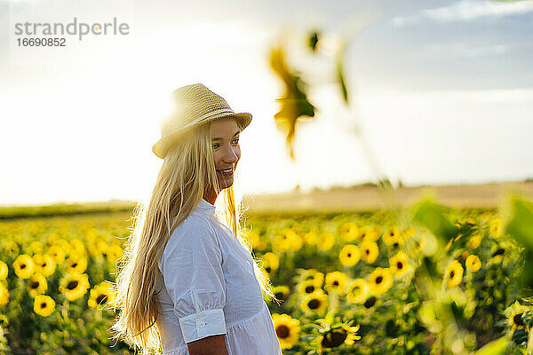 Junge attraktive blonde Frau posiert in ihrem Designerkleid in einem Sonnenblumenfeld und trägt einen Hut