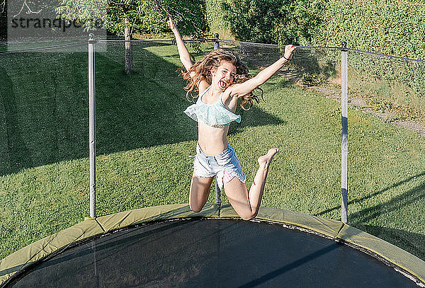 Horizontales Foto eines lächelnden jungen brünetten Teenagers  der auf einem Trampolin mit Netz auf dem grünen Hof im Freien springt. Das Mädchen trägt kurze Jeans und ein Top und sieht Spaß  aktiv