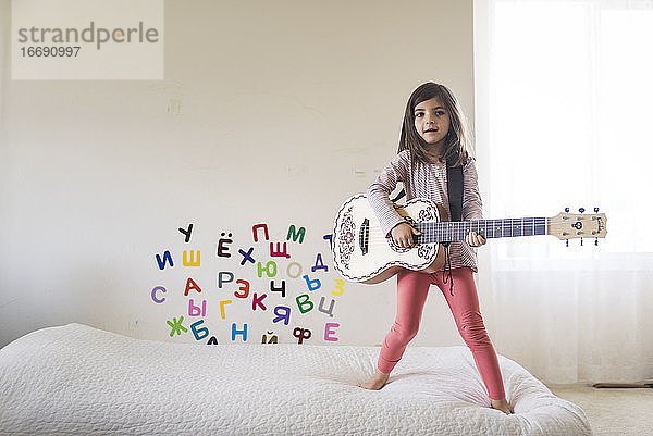 Junges Mädchen steht auf dem Bett und spielt Gitarre in einem Zimmer mit Buchstaben an der Wand